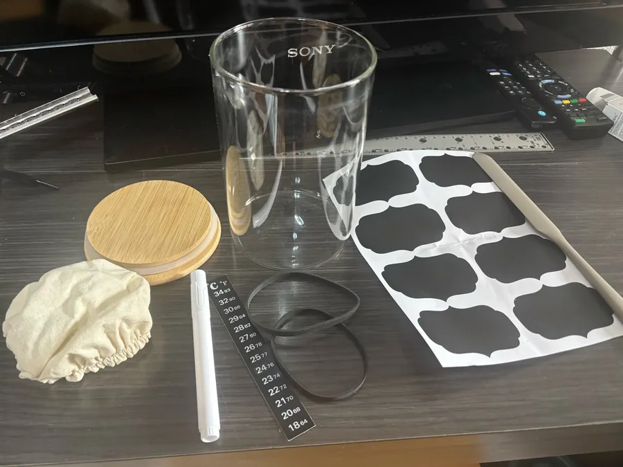 The full kit unpacked on a table: glass jar, wooden lid, cloth cover, spatula, stick-on thermometer and sheet of chalk labels